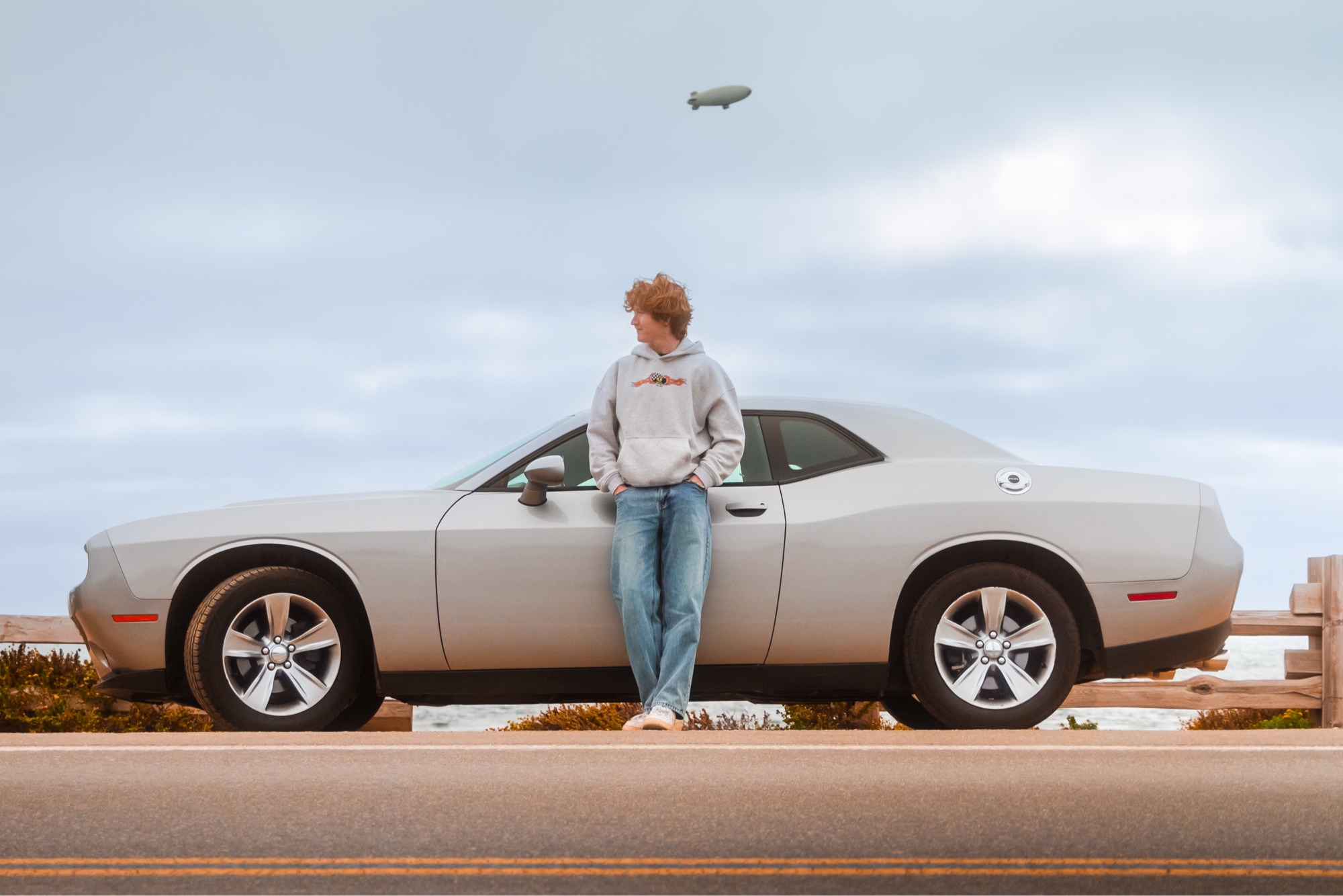 Person leaning on Dodge Challenger side, coastal highway, blimp overhead