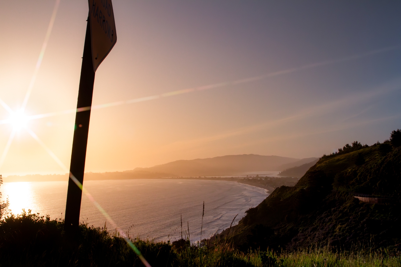 Coastal sunset over bay with signpost silhouette and sun flare