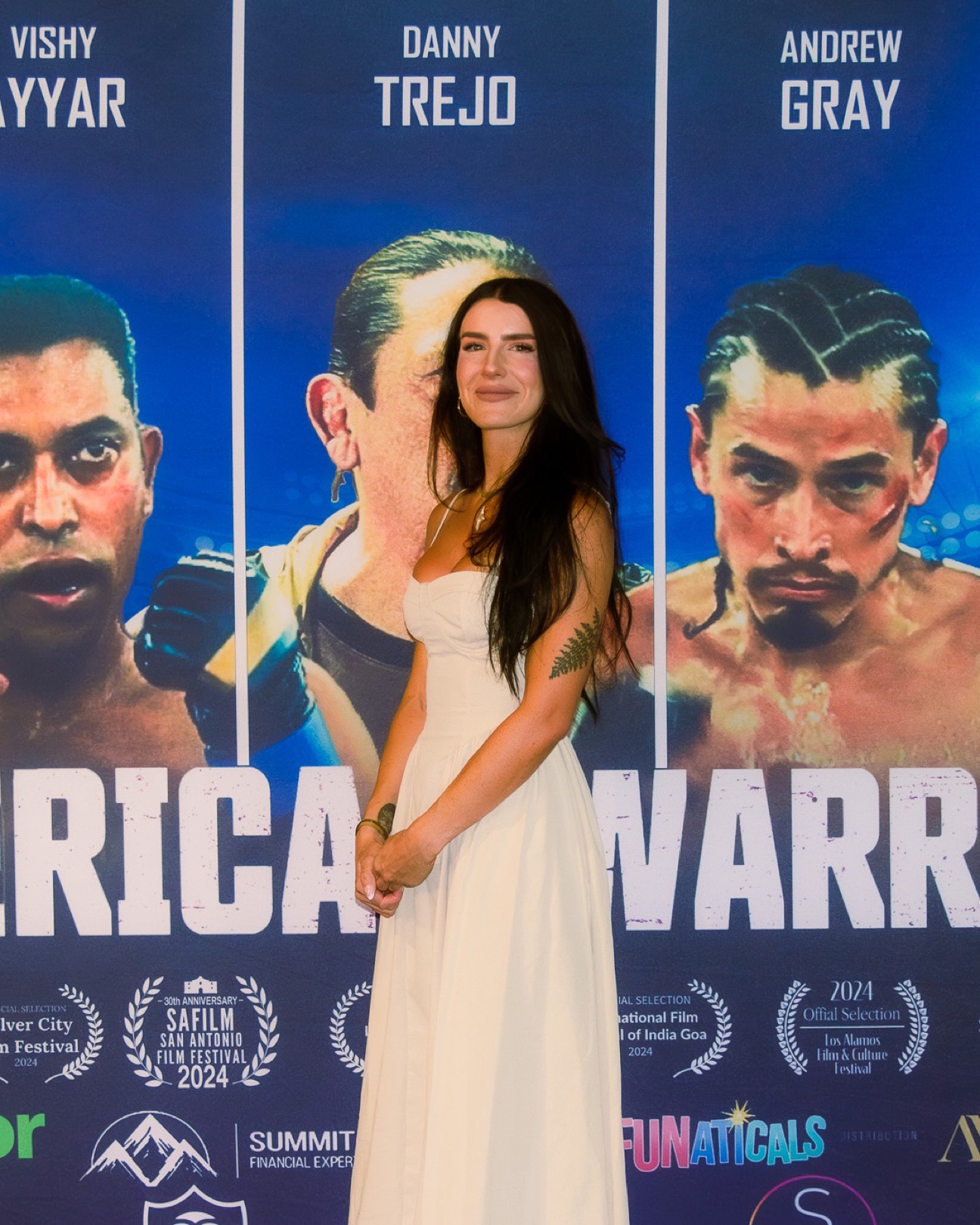 Woman in a long white sundress with dark hair over one shoulder, smiling softly with hands clasped at a film premiere