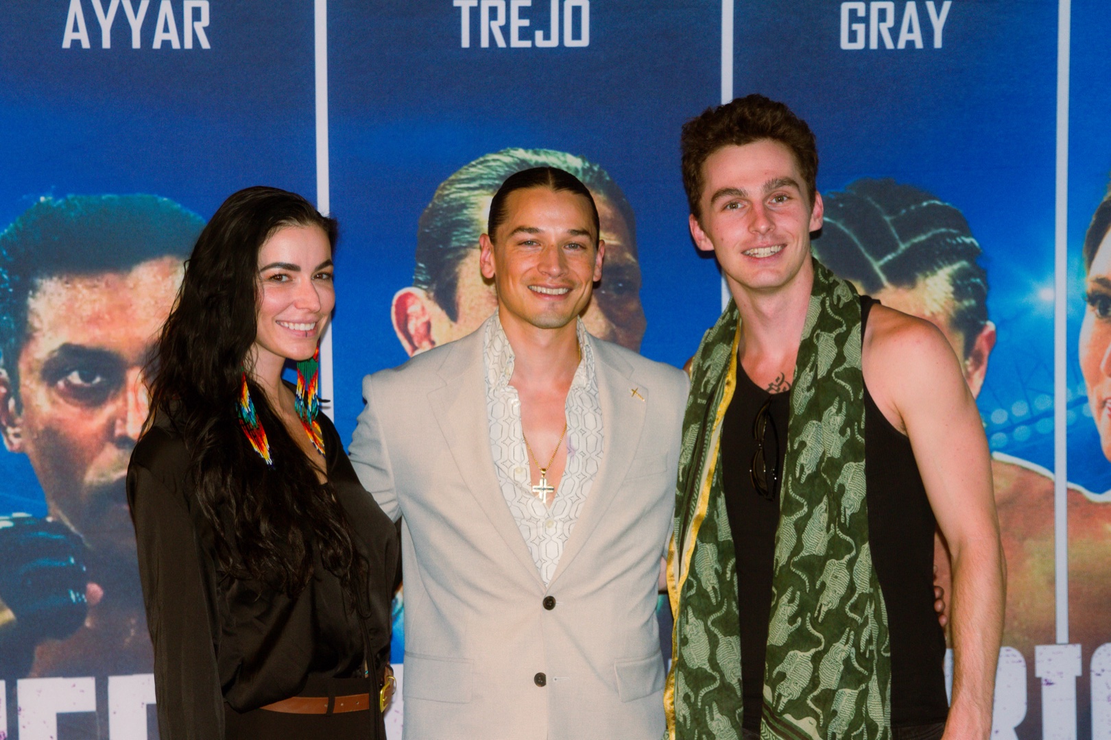 Woman in long beaded earrings, a man in a cream suit and a man in a green scarf smiling together at a film premiere
