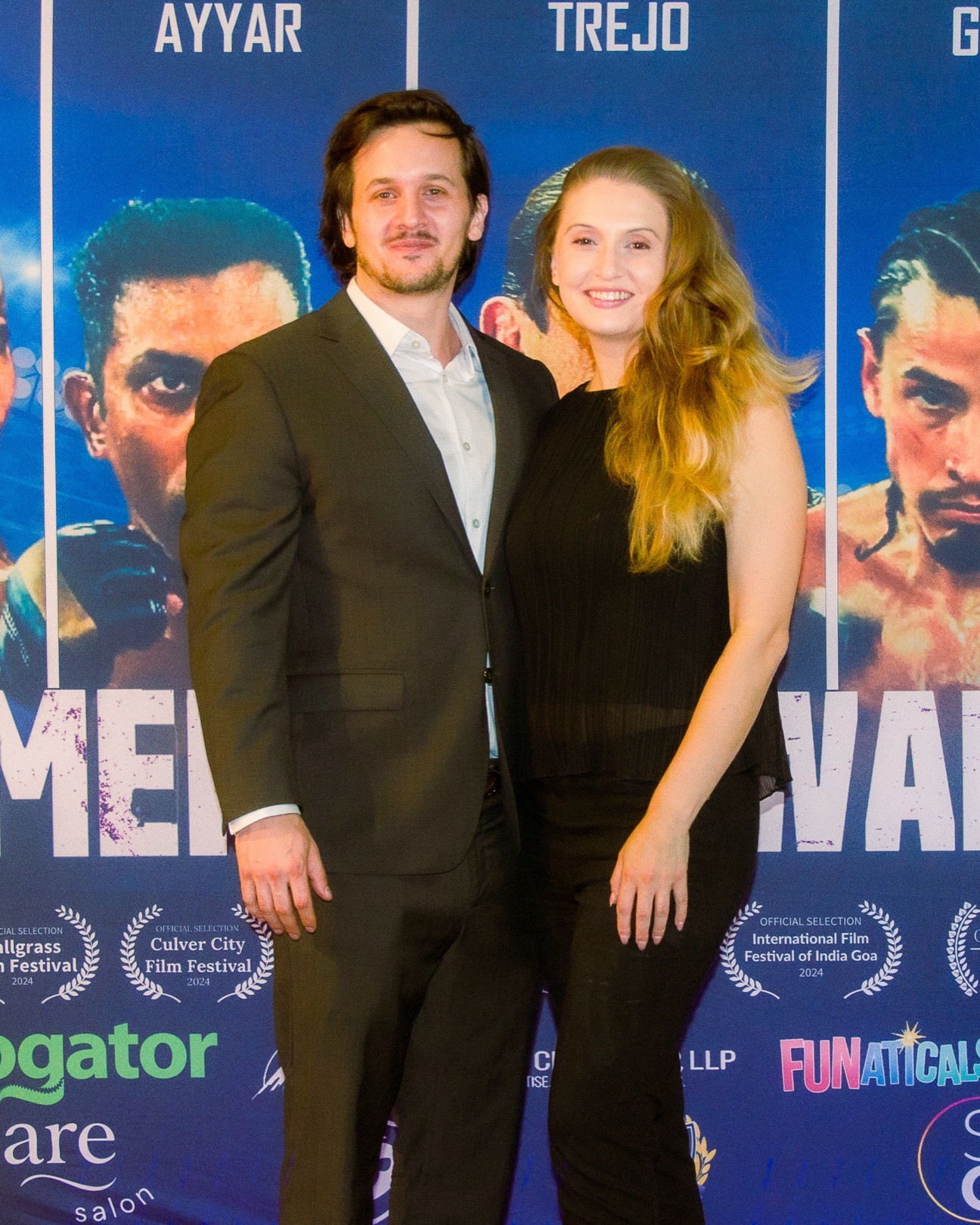 Couple at a film premiere, a man in a charcoal suit and open white collar beside a woman with long strawberry blonde hair