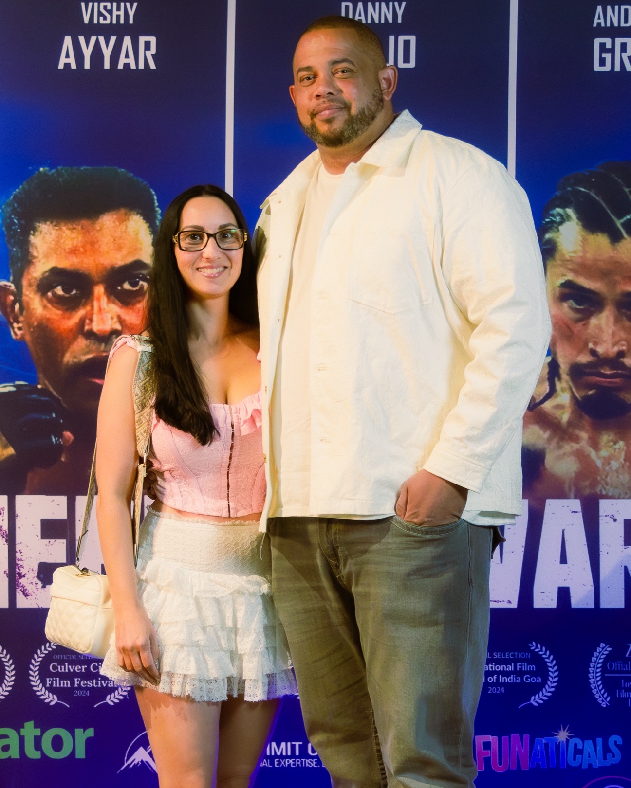 Couple at a film premiere, a woman in a pink corset top and ruffled white skirt beside a tall man in a cream overshirt