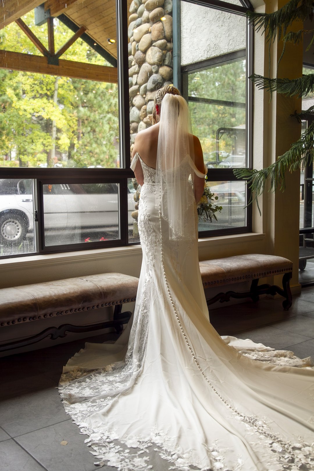 Bride seen from behind in a lace gown with a long train and veil beside tall lodge windows